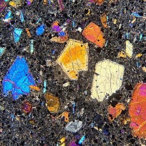 Könnte beinhalten: Eine mikroskopische Ansicht eines Dünnschliffs von magmatischem Gestein, das verschiedene Mineralkristalle mit leuchtenden Interferenzfarben wie Hellblau, Orange, Gelb und Magenta in einer dunklen, feinkörnigen kristallinen Matrix zeigt.