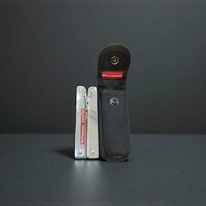 May include: A silver Victorinox SwissTool multi-tool with a black nylon carrying case. The multi-tool is partially open, revealing its various components. The case has a snap closure and a red Victorinox label.