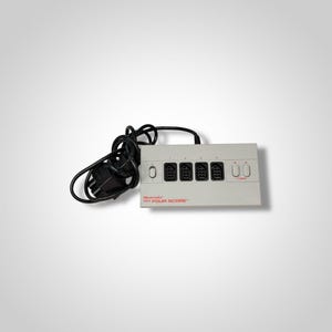 May include: A light gray Nintendo NES Four Score adapter with four controller ports and two turbo buttons. The adapter has a black power cord and is designed for use with the Nintendo Entertainment System.