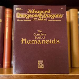 May include: A maroon-colored book titled "Advanced Dungeons & Dragons 2nd Edition" with gold lettering. The book is part of a collection of books on a wooden shelf. The title "The Complete Book of Humanoids" is also visible.