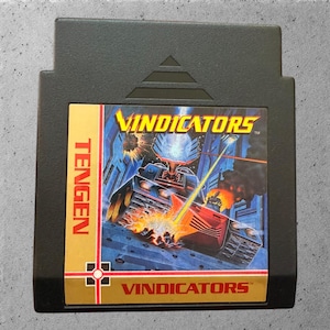 May include: Black Tengen Vindicators video game cartridge. The label features a futuristic tank firing a laser beam, with the word "Vindicators" in yellow. The Tengen logo is on the left side of the label.