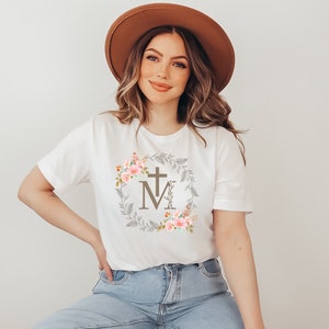 May include: White t-shirt with a floral wreath design featuring a cross and the letter "M" in the centre. The wreath is in shades of brown, pink, and green.