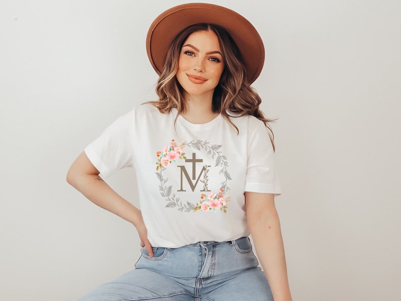 May include: White t-shirt with a floral wreath design featuring a cross and the letter "M" in the centre. The wreath is in shades of brown, pink, and green.