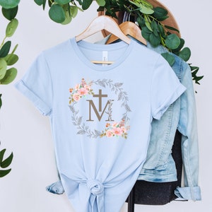 May include: Light blue t-shirt with a floral wreath design featuring a cross and the letters "M" and "M".
