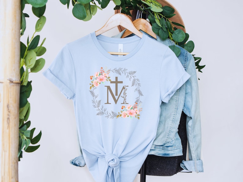 May include: Light blue t-shirt with a floral wreath design featuring a cross and the letters "M" and "M".