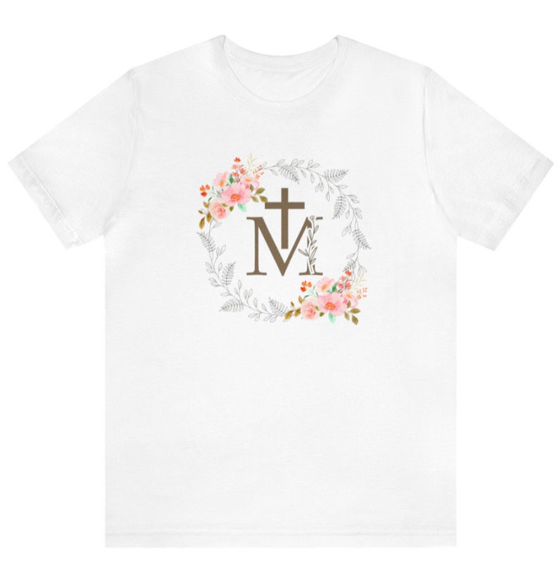 May include: White t-shirt with a floral wreath design featuring a brown cross and the letter "M".