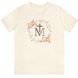 May include: A white t-shirt featuring a floral wreath design with a brown cross and the letter "M" in the centre.