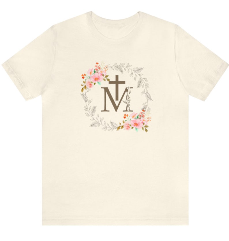 May include: A white t-shirt featuring a floral wreath design with a brown cross and the letter "M" in the centre.