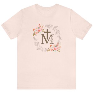 May include: A light pink t-shirt with a floral wreath design featuring a cross and the letter "M" in the centre.