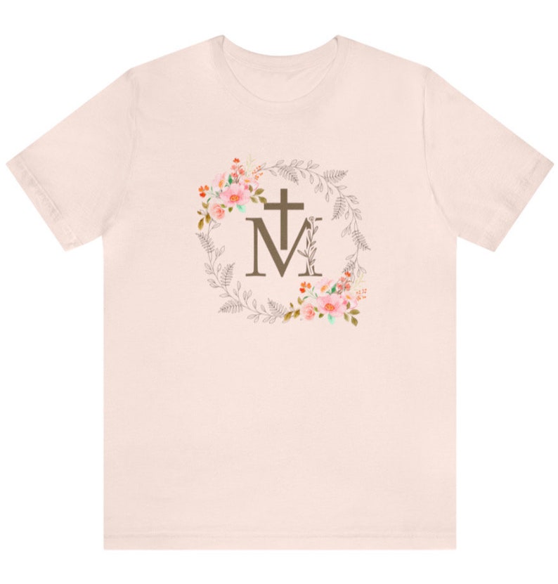 May include: A light pink t-shirt with a floral wreath design featuring a cross and the letter "M" in the centre.