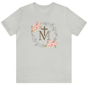 May include: A light grey t-shirt with a floral wreath design featuring a cross and the letter "M".