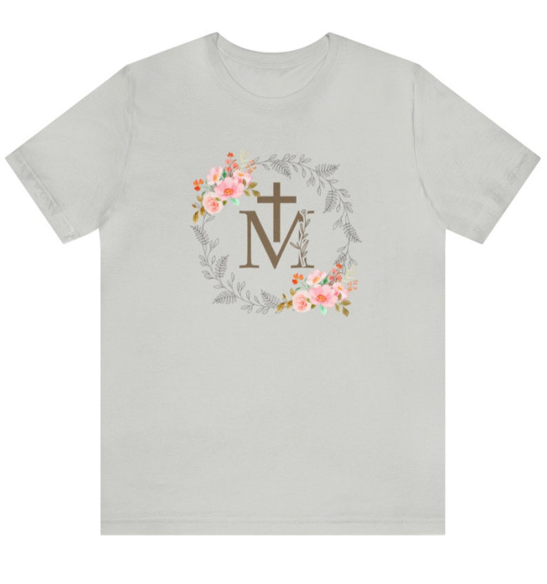 May include: A light grey t-shirt with a floral wreath design featuring a cross and the letter "M".