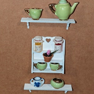 May include: A white shelf unit with three shelves. The top shelf has a green teapot and a green creamer. The middle shelf has two jars with labels that say "Belle du Jour" and a small potted plant. The bottom shelf has two green teacups and a blue and white teacup.