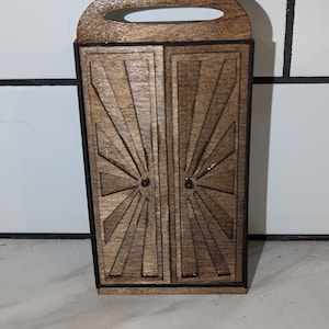 May include: A wooden box with a handle on top. The box has a decorative design on the front that resembles a sunburst. The box is brown and has a black border.