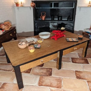 Può includere: Una scena da cucina in miniatura con un tavolo di legno preparato per la cottura. Il tavolo ha una ciotola di farina, un mattarello, una ciotola di uova, uno stampo per muffin e una bilancia rossa. Ci sono anche biscotti su un vassoio e una ciotola di frutta su un tavolo vicino.