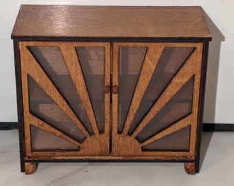 May include: A miniature wooden cabinet with a sunburst design on the doors. The cabinet is made of light brown wood with a darker brown trim. It has two doors with small wooden knobs and rests on small, curved feet.
