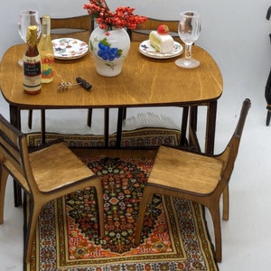 May include: A miniature wooden dining set with a brown table, two chairs, and a rug. The table is set with two wine glasses, a bottle of wine, a plate with a dessert, and a vase with red flowers.