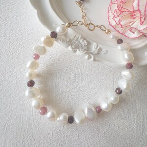 May include: A delicate bracelet made with white pearls and small, dark red gemstones. The bracelet has a gold clasp.