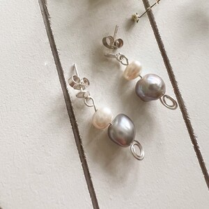 May include: A pair of silver earrings with a pearl and a grey pearl dangle. The earrings are on a white background.