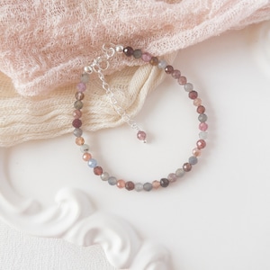 May include: A delicate bracelet made with small, faceted gemstones in shades of pink, blue, and grey. The bracelet is strung on a silver chain with a small silver charm.