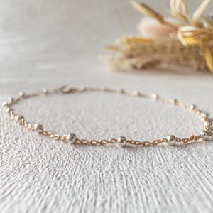 May include: A delicate gold chain bracelet with small silver beads. The bracelet is on a white textured surface.