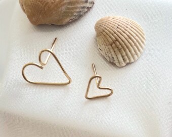 May include: Two gold heart-shaped earrings on a white fabric background. The earrings are different sizes.