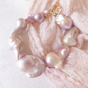 May include: A bracelet composed of large, irregularly shaped, pale pink pearls. The pearls are connected with a gold-coloured clasp and small accent pearls. The bracelet is displayed on a soft, pink, textured fabric.