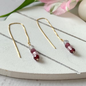 May include: A pair of gold chain earrings with three gemstone beads each. The beads are a gradient of light pink, dark pink, and burgundy.