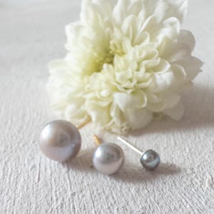 May include: Three pearl earrings of different sizes and shades, with a white flower in the background. The pearls vary from a large, light grey to a small, dark grey stud. The earrings are on a textured white surface.