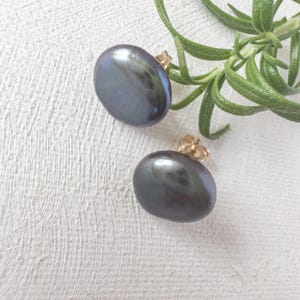 May include: Two dark grey oval pearl stud earrings with gold posts. The pearls have a lustrous sheen. They are set against a textured white background, with a sprig of green rosemary in the background.