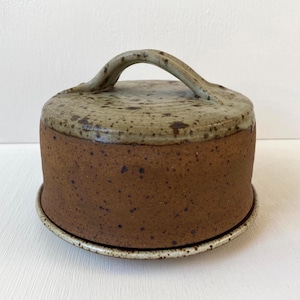 Pode incluir: Uma cúpula de queijo de cerâmica artesanal com uma alça. A cúpula é marrom com detalhes salpicados, e a tampa é de uma cor bege mais clara. A cúpula fica em um prato correspondente, perfeito para servir queijo ou outros aperitivos.