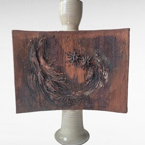 May include: A rectangular, textured brown ceramic plaque featuring abstract, raised swirling patterns and two star-like shapes. The plaque is mounted on a smooth, light beige cylindrical ceramic pedestal base with a flared bottom.