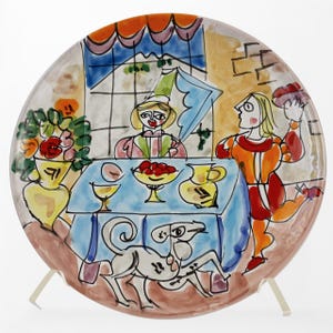 May include: A decorative ceramic plate featuring a colorful, whimsical scene. The artwork depicts figures at a table with food and a dog, with a window in the background. The plate has a brown rim and is propped up on a white stand.
