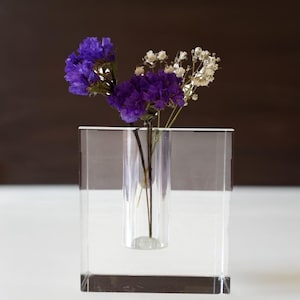 Può includere: Un vaso quadrato trasparente in acrilico con un inserto cilindrico che contiene fiori viola e bianchi. I fiori hanno piccoli e delicati fiori bianchi e vibranti grappoli viola. Il vaso è posto su una superficie bianca contro uno sfondo marrone scuro.