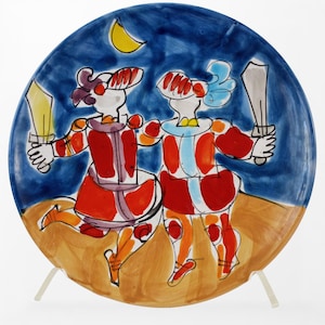 May include: A decorative ceramic plate featuring two stylized figures in red and white costumes, each holding a sword. The figures are set against a blue background with a yellow crescent moon. The plate is supported by a clear stand.