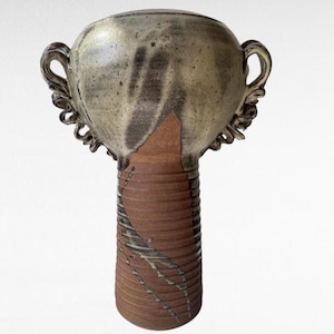 May include: A handcrafted ceramic vase with a unique design. The vase features a wide, rounded top with two ornate handles and a textured, cylindrical base. The color palette includes earthy tones of brown, tan, and hints of gray, creating a rustic aesthetic.