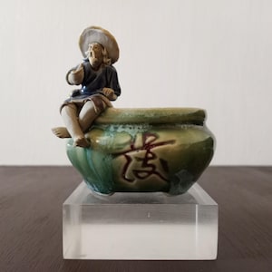 Op de afbeelding: Een groene en bruine keramische pot met een klein beeldje van een persoon die op de rand zit. Het beeldje draagt een blauw shirt en een strohoed. De pot heeft een Chinees karakter op de zijkant.