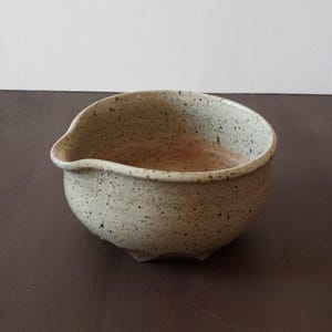 Puede incluir: Un cuenco de cerámica de color verde claro moteado con un pico vertedor. El cuenco tiene un fondo redondeado y un borde ligeramente acampanado.