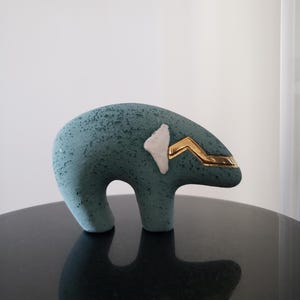 Könnte beinhalten: Eine türkisfarbene Bärenfigur mit strukturierter Oberfläche. Der Bär hat einen goldfarbenen Akzent auf dem Kopf und einen weißen Akzent an der Seite. Die Figur steht auf einer schwarzen, reflektierenden Oberfläche.