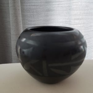 May include: A dark gray, round ceramic vase with a wide opening. The vase has a glossy finish and a subtle, geometric design near the base. The vase is sitting on a white surface.