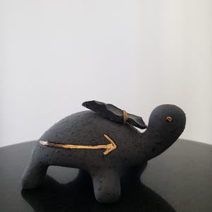 May include: A dark gray ceramic turtle figurine with a gold arrow pointing forward. A black, angular piece is attached to the turtle's back with a gold band. The turtle has a textured surface and a small gold dot for an eye.