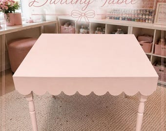 May include: A light pink square table featuring a scalloped apron edge and four slender, tapered legs. The table surface is smooth and flat, with the text "INTRODUCING The Darling Table" and "CRAFTED FOR CREATIVITY. MADE FOR MEMORIES." visible.