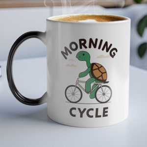May include: White ceramic coffee mug with a black handle. The mug has a green turtle riding a bicycle with the text "MORNING CYCLE" printed on it.