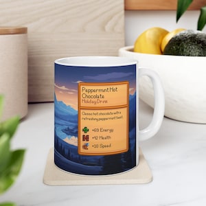 May include: White ceramic mug with a wintery landscape scene and a game-style text box that reads "Peppermint Hot Chocolate Holiday Drink" with stats: +69 Energy, +42 Health, +10 Speed.