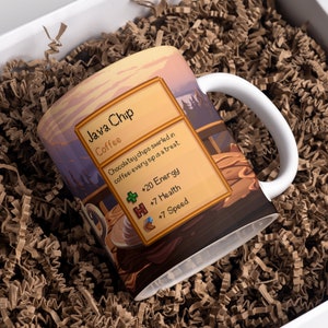 May include: A white ceramic mug with a brown handle. The mug has a colorful illustration of a coffee cup with chocolate chips and a game-style description that reads "Java Chip Coffee Chocolatey chips swirled in coffee every sip is a treat +20 Energy H +7 Health +7 Speed".