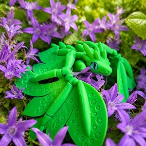 Puede incluir: Una figura de dragón articulada de color verde brillante con un diseño de alas similar a una hoja que presenta patrones circulares y un solo ojo azul. La criatura está colocada entre numerosas flores pequeñas de color púrpura en forma de estrella y follaje verde.
