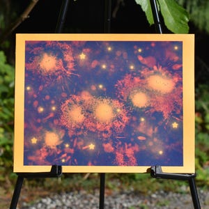 May include: A rectangular print featuring a dark blue background with glowing orange dandelion-like seed heads and scattered small yellow stars. The artwork is framed by a solid yellow border and displayed on a black easel stand.