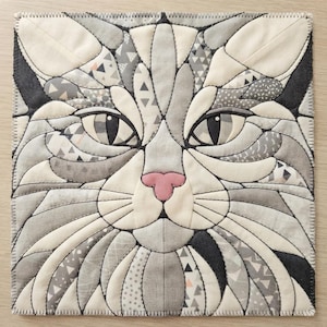 May include: A square fabric wall hanging featuring a stylized cat face composed of various gray, white, and black patterned fabric pieces. The cat has almond-shaped eyes, a small pink nose, and visible black stitching outlining each fabric segment.