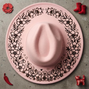 Op de afbeelding: Een lichtroze vilten fedora-hoed met een zwart inktachtig bloemenkransontwerp op de rand. Het ontwerp bevat bloemen, bladeren, kleine hartjes en een paar trouwringen. De hoed is omringd door kleine rode decoratieve voorwerpen.
