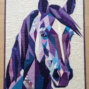 Könnte beinhalten: Ein rechteckiger Stoffquilt mit einem geometrischen Pferdeporträt. Das Design besteht aus verschiedenen violetten, blauen und hellrosa Stoffstücken mit sichtbaren Nähten, die auf einem cremefarbenen Hintergrund mit einem violetten Rand angeordnet sind.